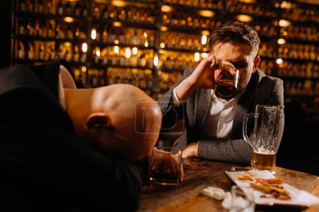 A young man in a suit sleeps near a glass of whiskey and beer on a table in a pub, another man drinks beer