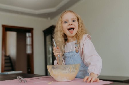 Photo for Little girl is cooking cookies in the kitchen, showing her tongue - Royalty Free Image