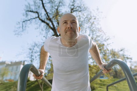 Foto de Calvo hombre en ropa deportiva está entrenando al aire libre con barras paralelas - Imagen libre de derechos