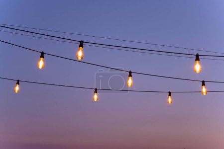Foto de Decoración festiva de bombillas amarillas. una cadena de luces brillan en una cena. Las bombillas seguidas sobre el alambre - Imagen libre de derechos