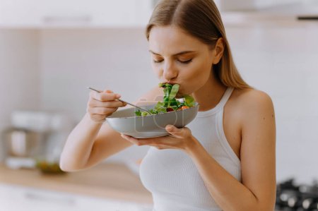 Téléchargez les photos : Jeune et heureuse femme mangeant une salade saine assise sur la table avec des ingrédients frais verts à l'intérieur. fille affamée mange une salade verte - en image libre de droit
