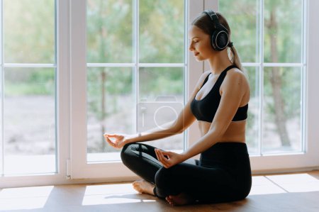 Foto de Hermosa y deportiva chica meditando y escuchando música en los auriculares en casa por la mañana. Su mañana junto a la ventana. posición de loto para la meditación - Imagen libre de derechos