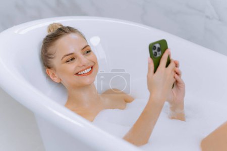 Téléchargez les photos : Portrait d'une belle femme tapant un message sur un téléphone portable tout en se baignant à la maison. Une femme dans le bain prend un selfie sur son téléphone. passer du temps dans le bain - en image libre de droit
