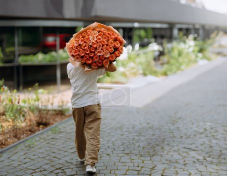 Foto de Un niño pequeño lleva un gran ramo de rosas a su novia. Niño con flores de rosa, atrás. camina detrás con flores - Imagen libre de derechos