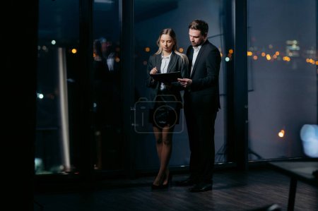Photo for A man and a woman are talking near the panoramic window on the background of the city. Work after work at night. male and female office workers talking. Business partners talking in night office - Royalty Free Image
