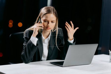 Angry woman with alarm clock talking by mobile phone in office at night. business woman working in the office