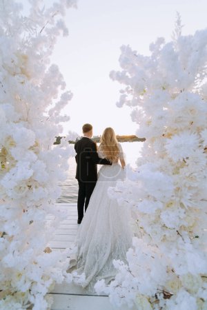 Téléchargez les photos : Couple de mariage près de l'arche avec des fleurs au coucher du soleil le jour du mariage. Un couple se tient debout, dos à dos, sur la rive de la rivière. vue arrière - en image libre de droit