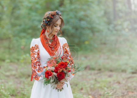 Une charmante mariée en robe rouge brodée tient un bouquet de mariage et pose dans un parc verdoyant. une fille en vêtements ukrainiens traditionnels dans la nature
