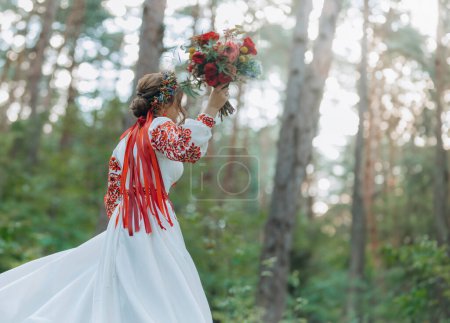 Eine bezaubernde Braut in einem rot bestickten Kleid hält einen Brautstrauß in der Hand und posiert in einem grünen Park. ein Mädchen in traditioneller ukrainischer Kleidung in der Natur