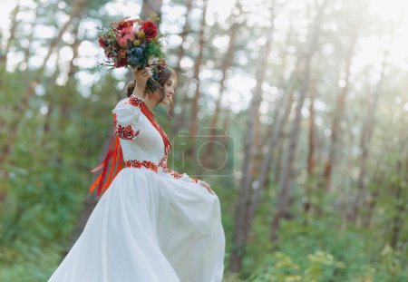 A charming bride in a red embroidered dress holds a wedding bouquet and poses in a green park. a girl in traditional Ukrainian clothes in nature