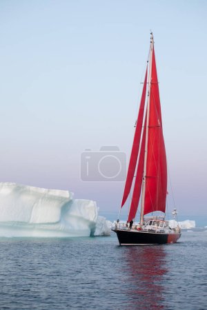 Segelboot mit roten Segeln, das bei Sonnenaufgang zwischen Eisbergen kreuzt. Disko Bay, Grönland.