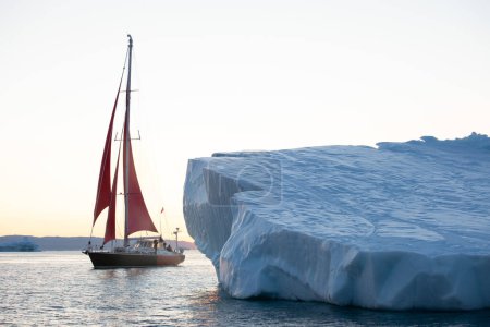 Sail boat with red sails cruising among icebergs during sunrise. Disko Bay, Greenland.