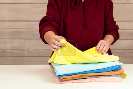 Téléchargez les photos : Une femme plie plusieurs t-shirts multicolores. Préparation des vêtements pour le don à la charité. Consommation consciente. Élimination des vêtements non utilisés. mode lent - en image libre de droit