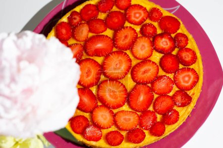 Foto de Homemade cheesecake garnished with strawberry slices whit table. Against the background of a bouquet of pink and yellow peonies. Sugar-free, low-carb summer dessert with sweetener. - Imagen libre de derechos