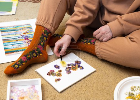 A middle-aged woman sits on a carpet and chooses pressed dried flowers for artwork. Hobbies for relax and enjoy of life.
