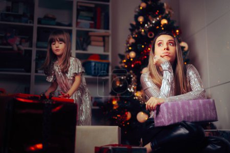 Téléchargez les photos : Maman débordée arrangeant des cadeaux sous l'arbre avec sa fille - en image libre de droit