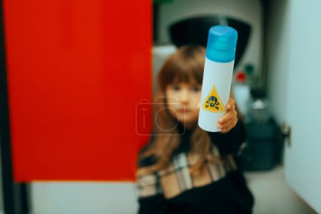 Téléchargez les photos : Little Girl Holding a Poisonous Substance from the Detergents Cabinet. Enfant en danger manipulant des sprays toxiques nocifs dans la cuisine - en image libre de droit