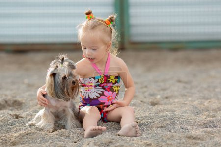 Téléchargez les photos : A little girl with Down syndrome sits on the beach with her dog. yorkshire terrier - en image libre de droit