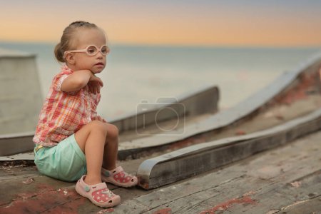 Téléchargez les photos : Little girl with Down syndrome sits on the summer coast - en image libre de droit