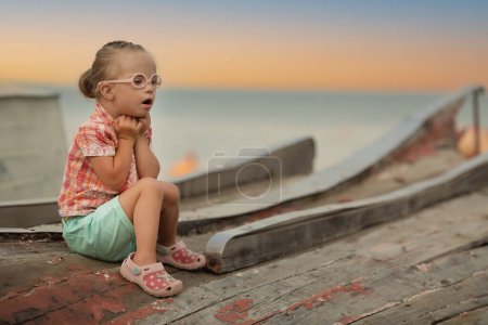 Téléchargez les photos : Little girl with Down syndrome sits on the summer coast - en image libre de droit