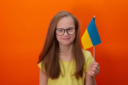 Foto de A girl with Down syndrome holds the flag of Ukraine. orange background - Imagen libre de derechos