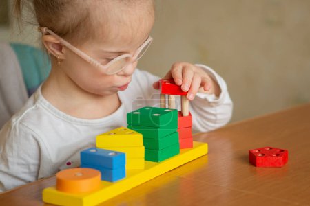 Photo for A beautiful little girl with Down syndrome learns to assemble an intricate pyramid. Multicolored geometric shapes - Royalty Free Image