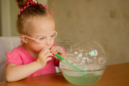 Téléchargez les photos : A beautiful little girl with Down syndrome learns to blow soap bubbles. Blow through a tube. Speech therapy exercises - en image libre de droit
