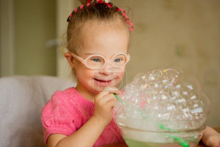 Téléchargez les photos : A beautiful little girl with Down syndrome learns to blow soap bubbles. Blow through a tube. Speech therapy exercises - en image libre de droit