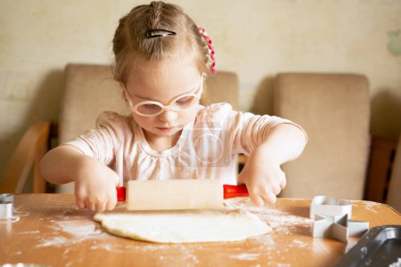 Téléchargez les photos : A girl with Down syndrome learns to roll out dough. Helps bake cookies in the kitchen - en image libre de droit