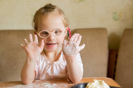Photo for A girl with Down syndrome learns to roll out dough. Helps bake cookies in the kitchen - Royalty Free Image
