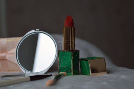 Foto de Cosméticos decorativos para niñas, lápiz labial rojo brillante y un pequeño espejo gris, una pequeña caja de regalo con un lazo sobre un fondo gris claro, un hueso de la ceja y un párpado - Imagen libre de derechos