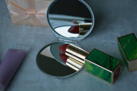 Foto de Cosméticos decorativos para niñas, lápiz labial rojo brillante y pequeño espejo gris, pequeña caja de regalo con lazo sobre fondo gris claro - Imagen libre de derechos
