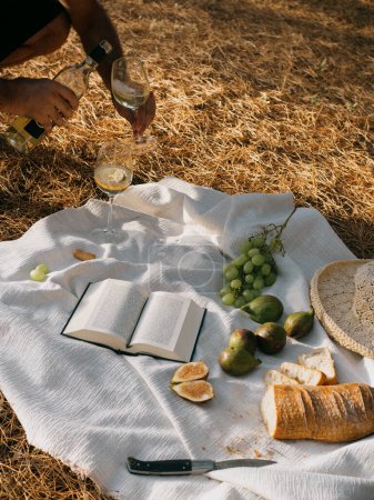 Photo for Man filling the wineglass. Vertical shot of a Mediterranean picnic for two in a pine forest. White Sardinian wine, open book, figs and grapes, cut bread, straw hat, white linen tablecloth on a soft carpet of pine needles - Royalty Free Image