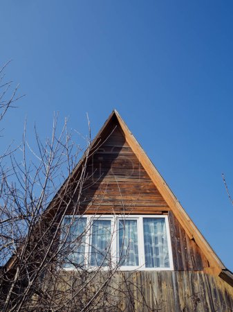 Vertical low angle shot of a wooden house rooftop with an attic window. Bare tree branches in the foreground and clear blue sky in the backdrop.