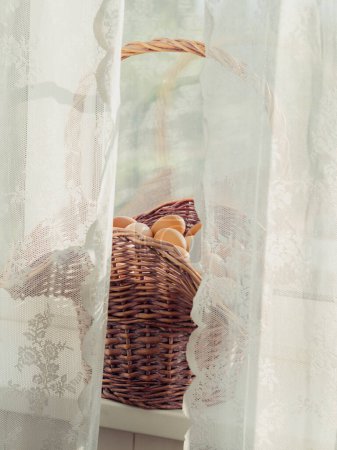 Vertikale Nahaufnahme eines Korbs voller Eier auf einem Fenstersims hinter weißen geschnürten Vorhängen.