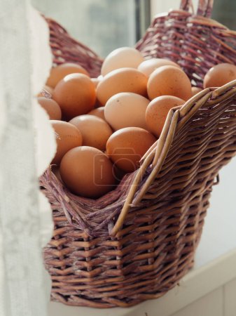 Vertikale Nahaufnahme eines Korbs voller Eier auf einem Fenstersims hinter weißen geschnürten Vorhängen.