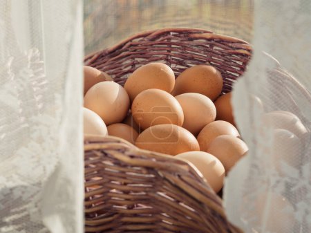 Horisontale Nahaufnahme eines Korbs voller Eier auf einem Fenstersims hinter weißen geschnürten Vorhängen.