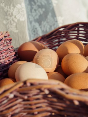 Vertikale Nahaufnahme eines Korbs voller Eier auf einem Fenstersims und weißer geschnürter Vorhänge im Hintergrund.
