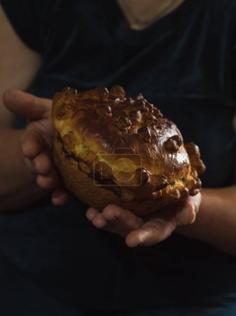 Vertical close-up of a woman's hands holding a traditional homemade sweet Ukrainian festive bread against a dark background. Bread slightly bent to one side.