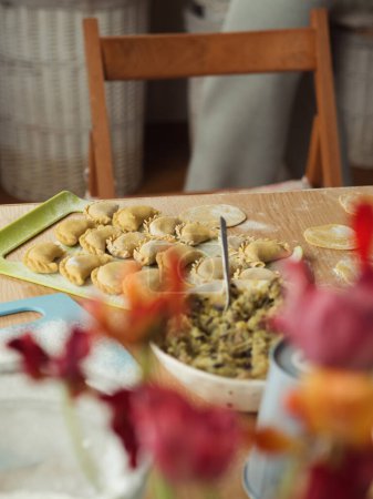 Acogedora cocina: Un primer plano vertical de la mesa con varenyky ucraniano en una tabla de cortar, tulipanes borrosos en primer plano, y una silla de cocina cerca. La harina se esparce por la mesa, un plato con relleno y una tabla de cortar vacía