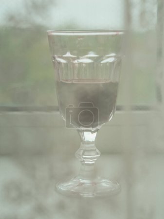 Vertical close up view of a glass of red wine on a windowsill behind a white laced curtain.