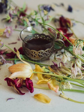 Flache Lage mit verblassten Blumen und einer Vintage-Kaffeetasse: Vertikale Nahaufnahme von verblassten Tulpen und Akeleienblüten auf einer weißen Oberfläche mit einer Vintage-Tasse schwarzen Kaffee in der Mitte.