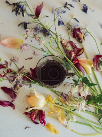 Flache Lage mit verblassten Blumen und einer Vintage-Kaffeetasse: Vertikale Nahaufnahme von verblassten Tulpen und Akeleienblüten auf einer weißen Oberfläche mit einer Vintage-Tasse schwarzen Kaffee in der Mitte.