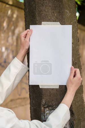 Foto de Las manos de las mujeres pegan una hoja de papel en blanco en un poste de luz. Burla. - Imagen libre de derechos
