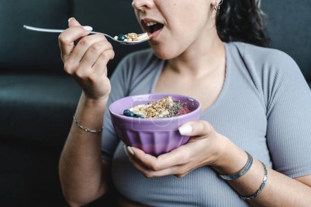 Hispanic young woman eating granola bowl with fruit and nuts at home. Healthy lifestyle and nutrition concept. Close up shot. Unrecognizable person