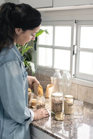 Hispanic woman re using recycled jars with bulk food in the kitchen. Plastic free and eco friendly concept. Vertical photography