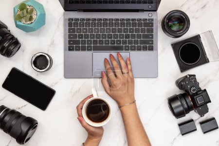 Téléchargez les photos : Top view of modern desk of woman photographer holding cup of coffee with laptop, camera, mobile phone and lenses on the table. Photography workspace and entrepreneur concept - en image libre de droit