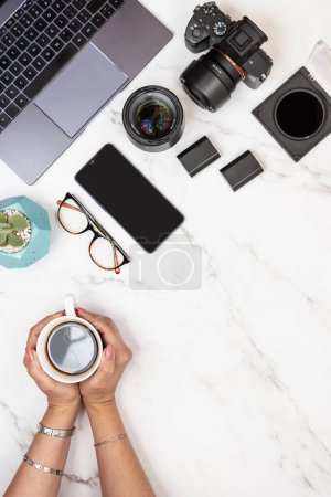 Photographer white marble desk with copy space on right holding coffee with hands. Vertical top view shot