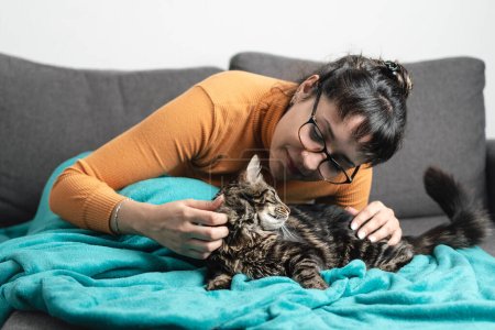 Latin woman petting her with love tabby cat while both are resting the sofa. Leisure activities and love for animals concept