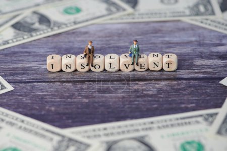 Foto de Dos pequeños empresarios en miniatura sentados en letras de madera de las que formaba la palabra insolvente, billetes de dólar alrededor - Imagen libre de derechos